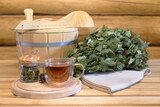Fototapeta Koty - A glass cup of herbal tea stands on a wooden bench next to a dry birch broom and traditional sauna accessories in the interior of a log bathhouse. 