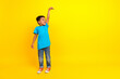 Full length photo of good mood child with curly hair dressed blue t-shirt look empty space show height isolated on yellow background