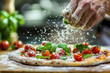 A chef applies the finishing touch to a pizza, dusting flour on top and garnishing with fresh tomatoes and basil, under the bright kitchen lights.