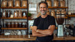 © Duka Mer - A man, wearing a black t-shirt and jeans, stands behind the counter at a coffee shop, arms crossed and smiling confidently.