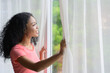 Happy young African female standing by bedroom window and opening sheer curtains at home