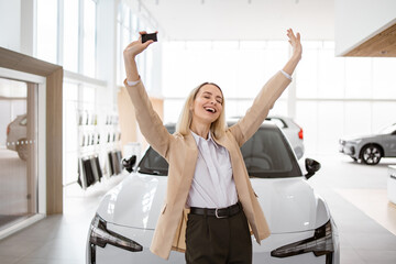 Cheerful woman standing near vehicle in dealership center and raising hands with excitement, copy space. Happy owner. Overjoyed Caucasian lady holding keys of new car after buying.