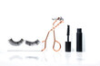 A several beauty products like fake eyelashes, tweezer and mascara on a white wall background