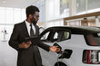 Young bearded African salesman, selling electric cars in light modern showroom with panoramic windows. Concept of buying eco-friendly car for progressive customer.