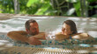 © MEDIAIMAG - Happy young lovers couple drinking sparkling wine while relaxing in the whirlpool bath hot tub at the spa outdoor pool in the exotic garden of luxury touristic resort in summer holiday