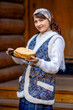 A young woman is dressed in national Russian dress. She is holding a plate of pancakes and a wicker basket in her hands. Greets guests for Maslenitsa.