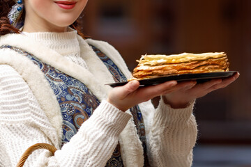 A young woman is dressed in national Russian dress. She is holding a plate of pancakes and a wicker basket in her hands. Greets guests for Maslenitsa.