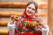 A beautiful Russian girl in red traditional Russian clothes holds bagels in her hands in the spring near a wooden house. Festive farewell to winter.