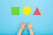 Baby boy hands and cut colorful geometric paper shapes on light blue table background. Pastel color. Time to learning. Infant development. Closeup. Point of view shot. Top down view.