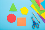 Fototapeta Tulipany - Colorful geometric shapes, scissors, pencil and application paper on light blue table background. Pastel color. Closeup. Child made different forms for learning. Top down view.