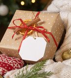 Fototapeta Boho - Gift tag on a Christmas present near cosy heart and sweater against Christmas tree lights, mockup