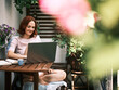 A cheerful woman sits with a laptop on a wooden table amidst a vibrant display of balcony greenery