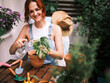 Woman in casual overalls waters a potted houseplant, surrounded by an abundance of balcony greenery