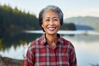 Portrait of a grinning asian woman in her 50s wearing a comfy flannel shirt isolated in serene lakeside view