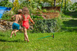Two happy kids in summer run around sprinkler at their backyard