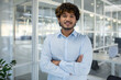 © Liubomir - A portrait of a confident young businessman with curly hair, standing arms crossed in a light-filled, contemporary office environment.