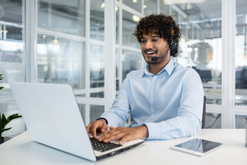 Wall Mural - Cheerful male customer service representative with headset working on laptop in a bright, contemporary office setting. Professional communication and support.