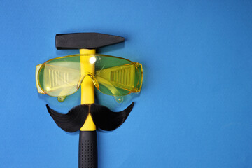 Sticker - Man's face made of artificial mustache, safety glasses and hammer on blue background, top view. Space for text