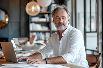 Wall Mural - Photo of middle aged man working on laptop in modern office, sitting at desk with papers and glass windows behind him looking into camera, shot from front