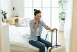 Fototapeta  - Young woman with a suitcase sitting on bed in hotel room and using phone
