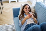 Fototapeta  - Smiling woman using mobile phone on sofa in living room at home
