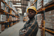 A confident black man in a warehouse wears a helmet, showcasing professionalism and expertise in the industrial sector.