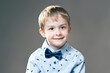 Portrait of little boy with brown eyes in blue shirt and bowtie looking straight into the camera and smiling, gray background