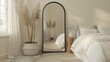 an elegant bedroom adorned with a large black arch mirror, accompanied by a small white wicker basket filled with items and a modern floor lamp styled after bamboo.