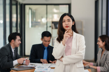 Confident young businesswoman standing in office with colleagues discussing in background. Leadership and career concept