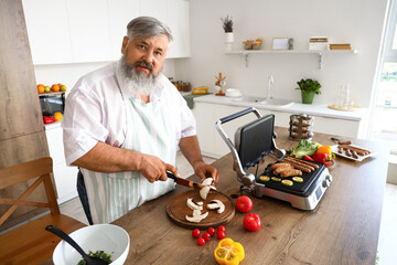 Canvas Print - Mature man cooking tasty sausages on modern electric grill and cutting fresh vegetables at table in kitchen