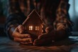 Close-up of hands cradling a warmly lit miniature house model in the dark.