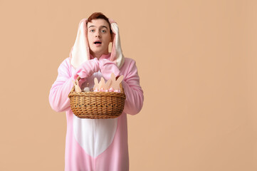 Wall Mural - Surprised young man in Easter bunny costume holding basket on beige background