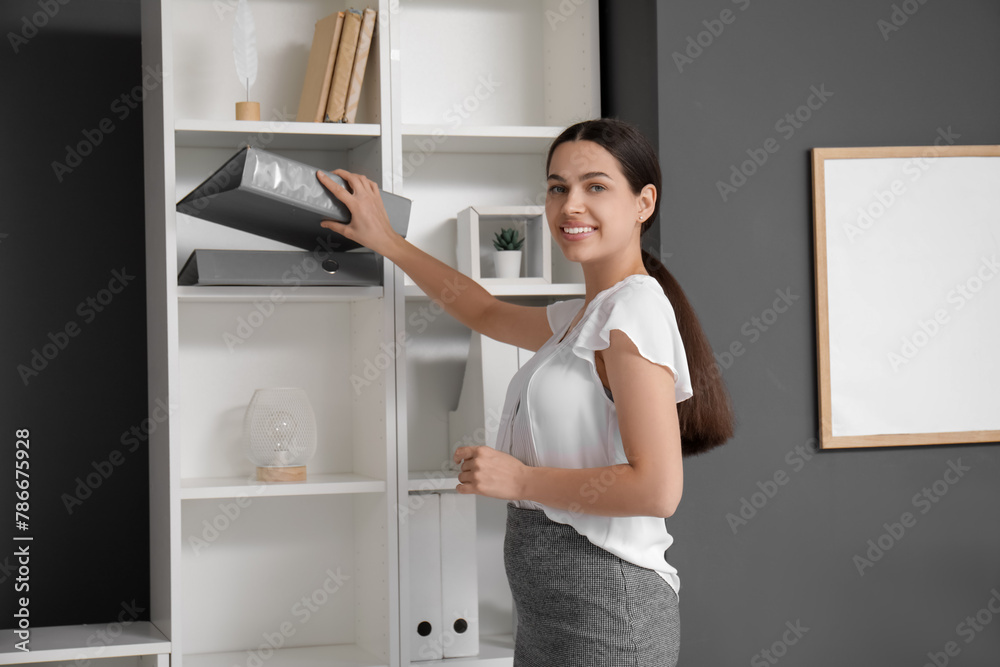 Pretty businesswoman putting folder in shelving unit in light office