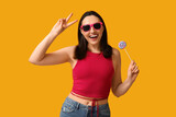 Fototapeta Tulipany - Beautiful young woman in sunglasses with sweet lollipop showing victory gesture on yellow background