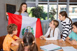 Excited young female teacher showing flag of Peru to schoolchildren preteens during history lesson in classroom