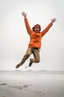 Person in vibrant orange jacket and red beanie joyfully jumping on a sandy beach with a foggy overcast sky.