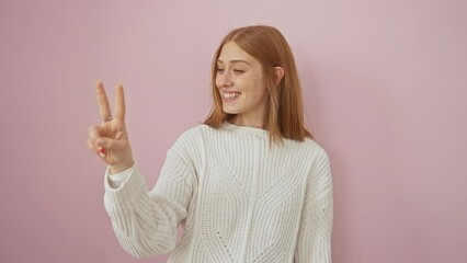 Canvas Print - Cheerful young redhead woman standing confidently, raising two fingers, and smiling. she's wearing a sweater, pointing up, showing number 2 over an isolated pink background