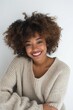 A cheerful young woman with curly hair wearing a cozy sweater flashes a bright smile against a white background.