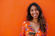 © AI_images - A vibrant Indian woman, in her late 20s, smiles warmly while recommending a smartphone app on a vibrant orange background, demonstrating her tech-savvy nature and infectious energy.