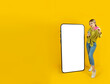 Cheerful blonde 20s girl showing credit card, standing near big huge mobile cell phone with empty blank touch screen mock up. Yellow studio background, copy space. Banner advertisement design concept.