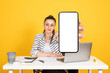 Show blank screen mobile phone mock up, young smiling positive employee businesswoman holding big empty display smartphone close up to camera. Sit office desk use laptop. Achievement career concept.
