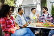 Cheerful multinational company having fun at a picnic. Multisexual friends sitting at outdoor table, celebrating food and drinking drinks. Two men shake hands.