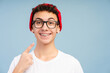 Smiling teenage boy with braces wearing stylish red hat and eyeglasses, pointing finger at his teeth