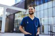 A male nurse is standing outdoors wearing scrubs and a stethoscope. He is smiling and looking at the camera. Portrait of male nurse. Portrait of mid adult nurse man at hospital