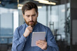 © Tetiana - Close-up photo of a young man in a blue shirt who is seriously sitting at the desk in the office and thoughtfully looking at the screen of the tablet he is holding in his hands