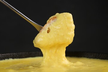 Dipping piece of bread into fondue pot with tasty melted cheese against dark gray background, closeup