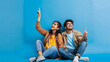 © Generative AI - Loving cheerful young indian couple wearing casual outfits sitting on floor looking up and pointing at copy space above for advertisement on blue studio background full length Great of : Generative AI