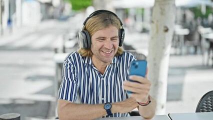 Sticker - A cheerful man using headphones video calls on his smartphone at an outdoor cafe table.