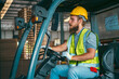 © chokniti - A man in a yellow safety vest and a hard hat is sitting in a forklift cab. He is holding a walkie talkie and smiling, foreman job to working in industrial factory storage store of global business
