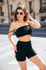 Wall Mural - Young beautiful smiling female in trendy summer black cycling shorts and tank top clothes. Sexy carefree woman posing in the street. Positive model. Cheerful and happy.  In sunglasses